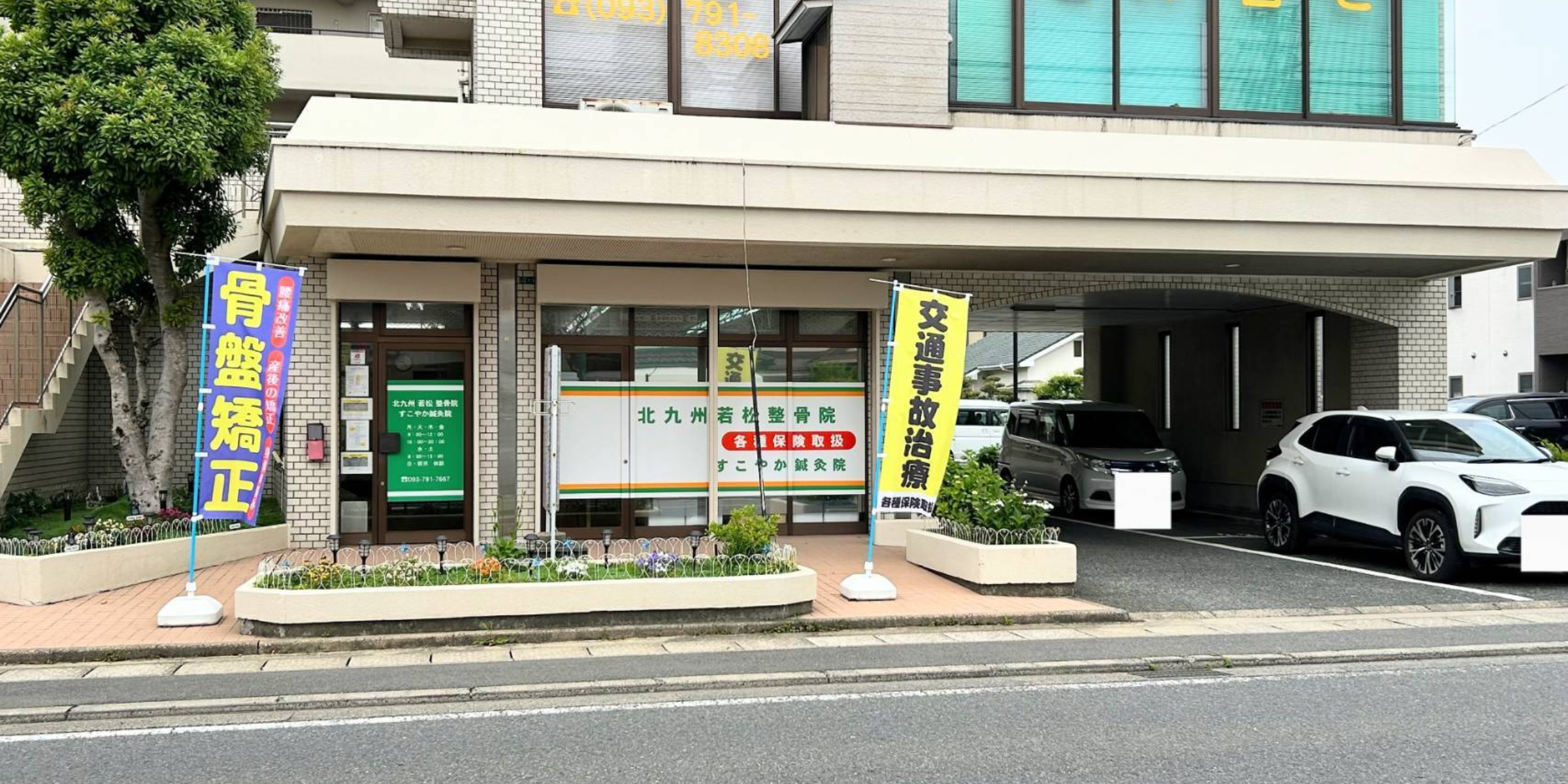 北九州市若松区の「すこやか鍼灸整骨院」の外観。入口には「骨盤矯正」「交通事故治療」ののぼり旗が掲げられており、窓には各種保険取扱の案内が表示されている。