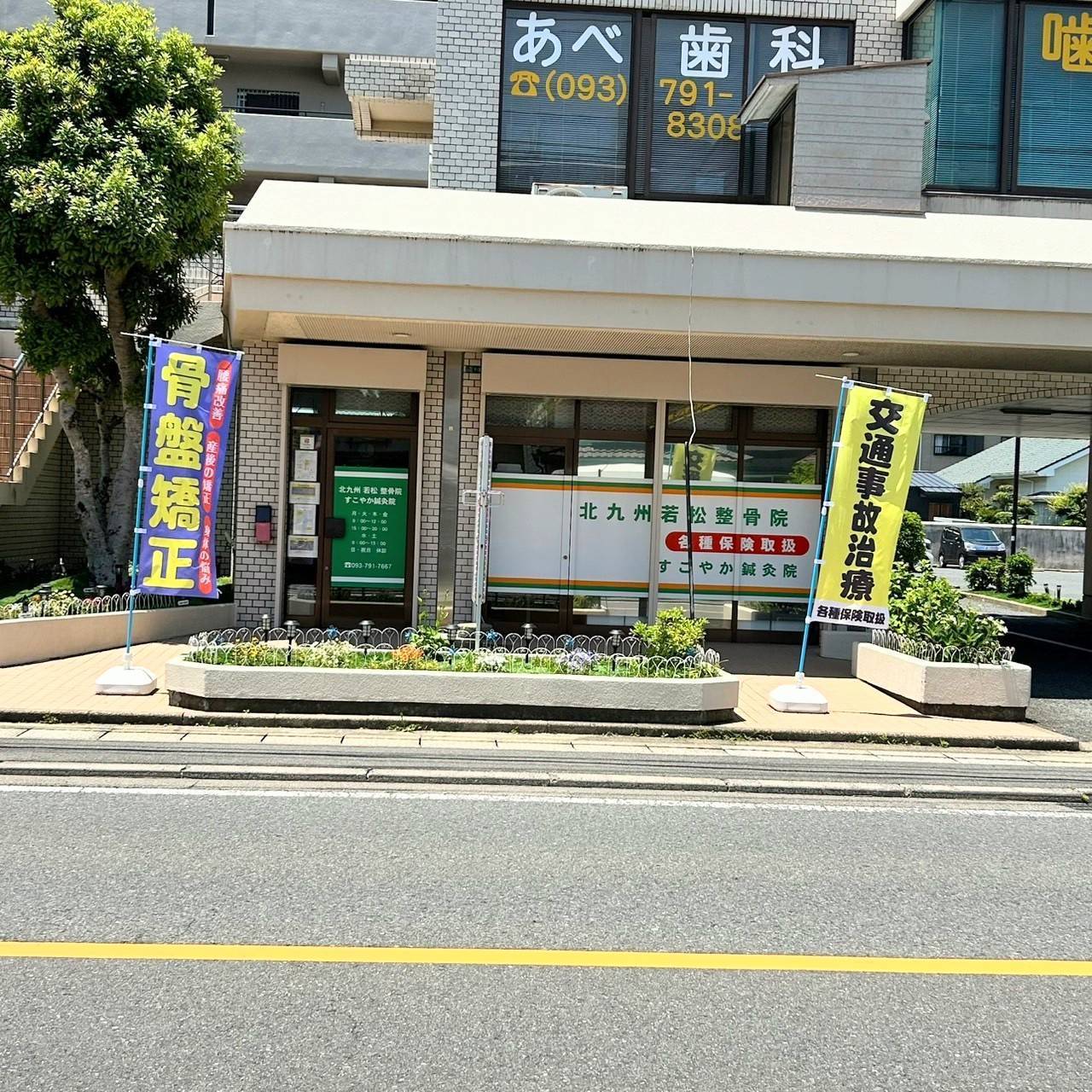 北九州市若松区の「すこやか鍼灸整骨院」の外観。入口には「骨盤矯正」「交通事故治療」ののぼり旗が掲げられており、窓には各種保険取扱の案内が表示されている。